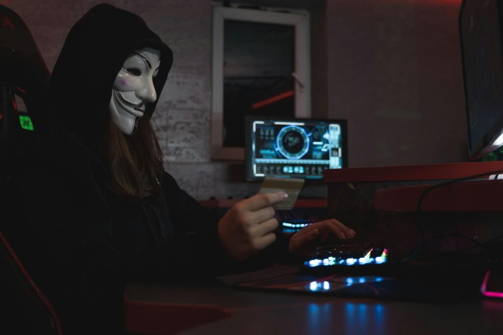 Masked hacker with credit card at computer, symbolizing cybercrime and anonymity.